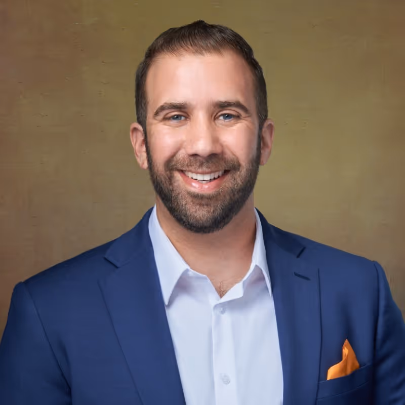 Smiling man with short dark hair and beard wearing a blue suit jacket, white shirt, and orange pocket square against a brown background.