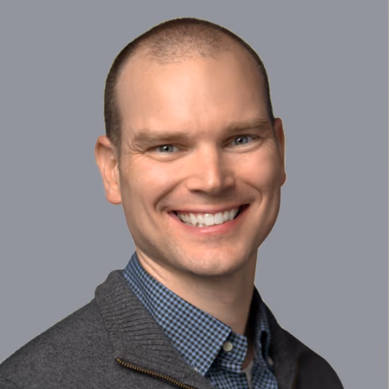 Smiling bald man wearing a gray sweater over a blue checkered shirt against a gray background.