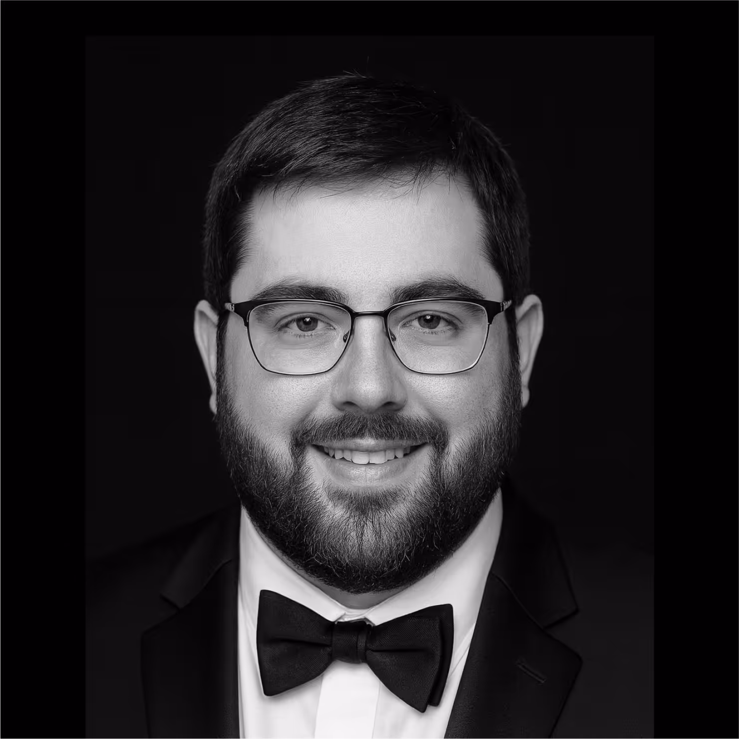 Smiling man with glasses and beard wearing a tuxedo and bow tie against a black background.
