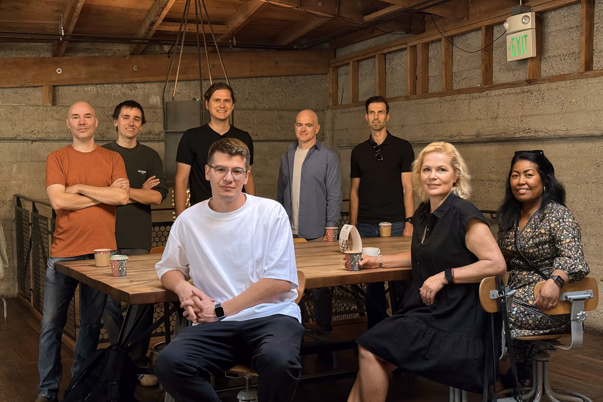 Group of eight adults, five standing behind and three seated around a wooden table in a rustic room with concrete walls and wooden ceiling.