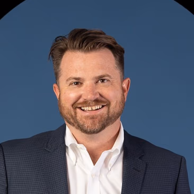 Smiling man with short brown hair and beard wearing a white shirt and dark blazer against a blue background.