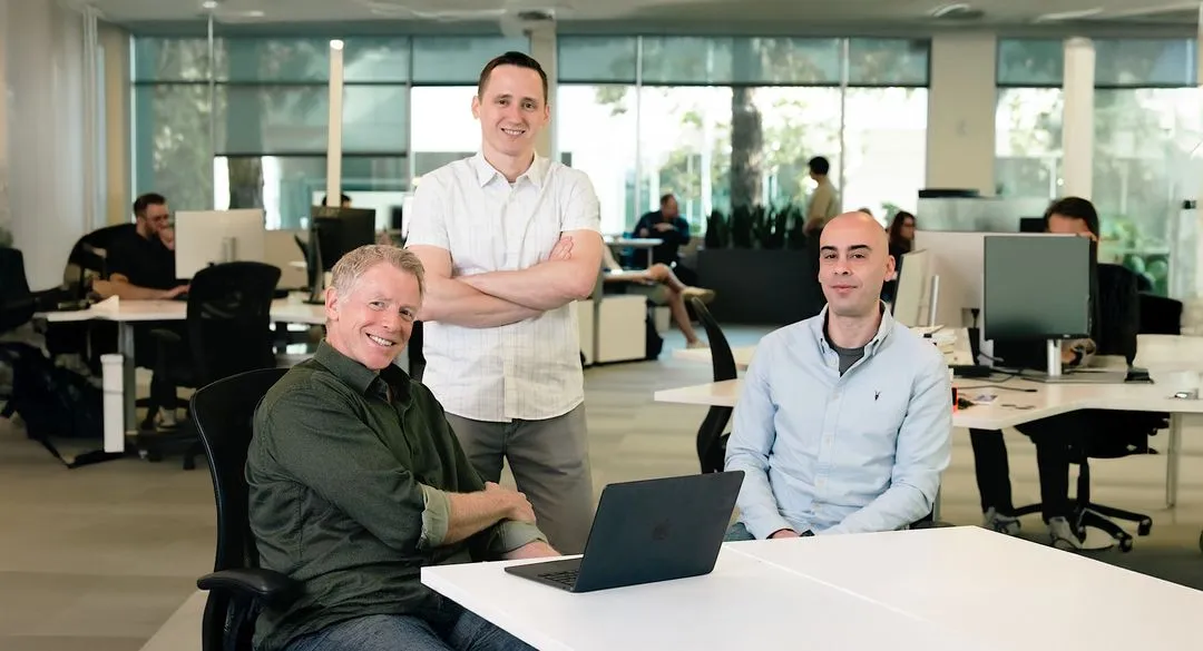 Three members of Augment Code's leadership team seated and standing around a laptop in the company's open-plan Palo Alto office