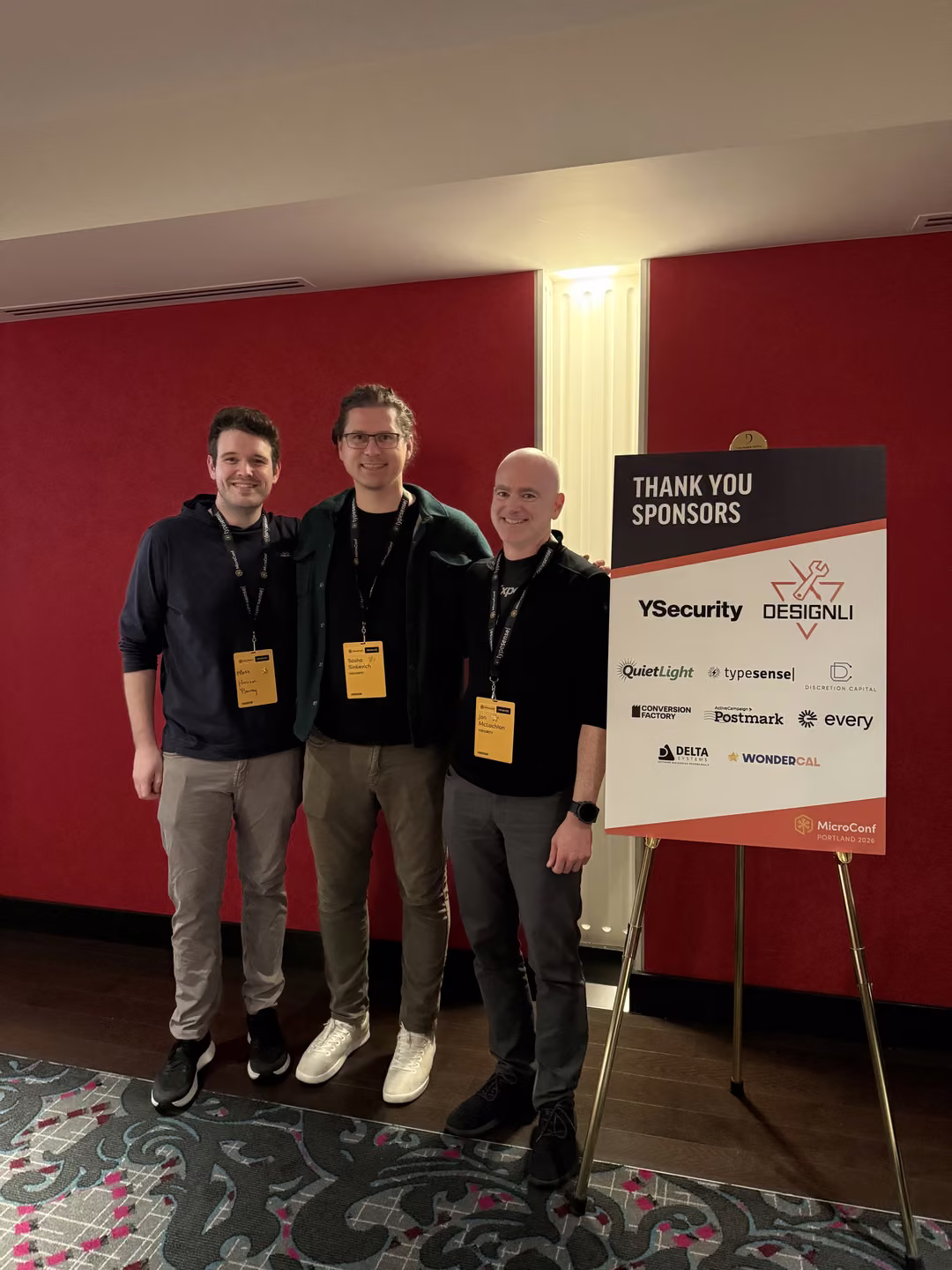 Three members of the YSecurity team beside the MicroConf US 2026 sponsor board listing YSecurity