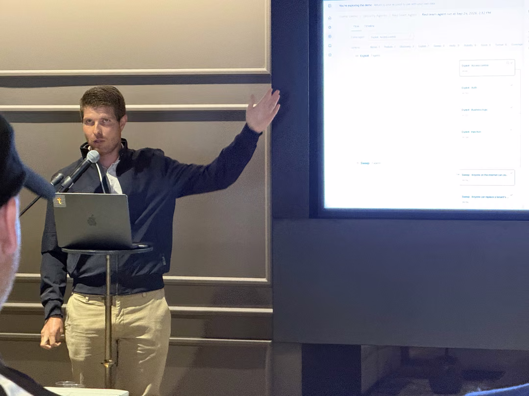 A presenter at a laptop podium gesturing to a live demo of an autonomous red team agent on a large screen at Tolmo Day in San Francisco