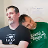 Two men sitting close together, one resting his head on the other's shoulder, both wearing black T-shirts with white text.